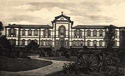 A monochrome photograph showing a path winding through a tropical garden, and leading to a large, two-story neoclassical building with tiled roof, walls in white stucco with dark stone used for quoin blocks, window frames and balustrades
