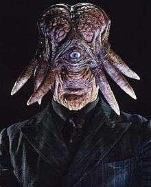 An alien creature with a vertically elongated head, small mouth, prominent chin and large exposed brain. A single grey eye is set centrally in the face, with tentacles protruding from the sides of the head. It wears a black shirt, tie and black pinstripe jacket.
