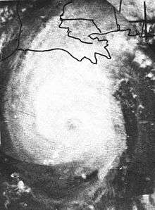 Black and white image of a slightly elongated tropical cyclone with a faintly visible eye at its center. Clouds appear white, while landmasses and bodies of water appear in darker shades of gray.