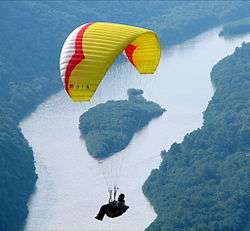 A person dangles below a red, yellow and white parasail, in the background is a lush forest with a large river and a tree-covered island.