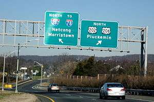 A multilane highway in a suburban area at a split, with two green signs over the road. The sign on the left reads north Interstate 287 to Interstate 78 Netcong Morristown with an arrow pointing to the upper right and the sign on the right reading north U.S. Route 202/U.S. Route 206 Pluckemin.