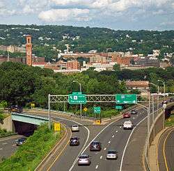 At bottom, one roadway of an expressway divides as it approaches a city skyline above it in the distance with the clock tower dominant on the left. Behind the city is a wooded hillside with development interspersed. Traffic on the road is heading away from the camera, following a left off ramp with a sign above it directing traffic to a place called Torrington via a highway with the number 8 in black on a white square; while the two main lanes veer right following a highway numbered "84" to the east in white on a semicircular red and blue background with white trim.