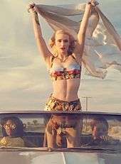 A portrait of a young blonde woman standing upright in a convertible car that is driving through a desert environment. She is waving a large piece of fabric material in the wind while two of her female friends are in the front seats of the convertible. The woman sports slick dark red lipstick, a tropical print bra-let and shorts.