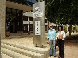 The entrance to the IRCAM (Institut Royal de la Culture Amazighe) building in Rabat. Two women are standing in front of a large, metal plaque with the word "IRCAM" written on it in large letters, and the organization's logo.