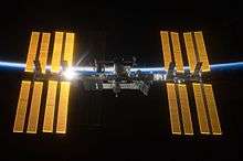 An image of the International Space Station. The silver-colored center module is dark blue, surrounded by four golden solar arrays on each side. The sun is reflecting off of the set to the left. In the background is the outline of the Earth.