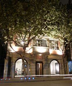 photo of a colonial style building lit up at night. The first two floors of the building are visible, with the top floors obscured by trees with dense foliage. The interior of the gallery can be viewed from outside through two ceiling to floor windows.