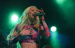 A young blond woman singing into a microphone on stage. She is wearing a colourful top and poses with her right hand on her chest as green stage lights shine upon her.