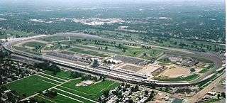 Aerial view of a rounded rectangular raceway that is partially surrounded by grandstands; within the racetrack is a pagoda and golf course