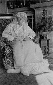 A monochrome portrait photograph of an elderly woman sitting in her home with a white Persian cat resting at her feet. The woman is wearing a light-colored dressing gown, her closed hands on her lap, her head tilting to one side, looking tight-lipped at the photographer, a white lace veil and scarf over her hair and framing her face.