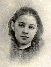 A soft photographic portrait of a girl approximately 11 years old, shown from the neck up, wearing a simple dark garment with no collar, her hair parted in the middle and falling straight to frame her cheeks, turning to ringlets at her neck and shoulders, the head tilted slightly to the right, the eyes looking directly forward. The image shows loss of detail from wear.