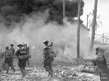 A black and white photograph showing soldiers advancing across a beach through smoke and debris