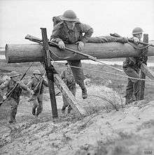 Two soldiers clamber over a wooded obstacle. Two more soldiers, in the background, run towards the obstacle.