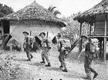 Soldiers wearing slouch hats and carrying rifles march past huts in a jungle clearing