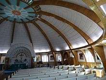Wooden pews under a domed white ceiling with regular wooden vaults. Paintings and windows alternate along the walls
