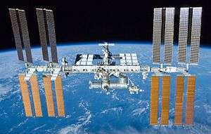 A rearward view of the International Space Station backdropped by the limb of the Earth. In view are the station's four large, gold-coloured solar array wings, two on either side of the station, mounted to a central truss structure. Further along the truss are six large, white radiators, three next to each pair of arrays. In between the solar arrays and radiators is a cluster of pressurised modules arranged in an elongated T shape, also attached to the truss. A set of blue solar arrays are mounted to the module at the aft end of the cluster.
