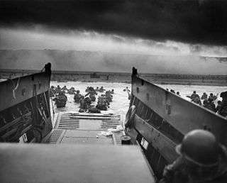 A black and white image of troops leaving an amphibious troop transport. They are walking through the water to a beach.