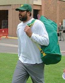 A beard man wearing a green cricket cap and a white shirt with a bag on his back.