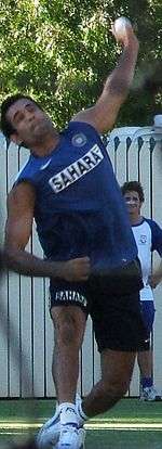 Young brown skinned clean shaven man wearing a blue sleeveless T-shirt with the word "SAHARA" in black, wearing dark shorts is about to deliver a white cricket ball. His left bowling arm is above his head and he is wearing white sports shoes. His leading right leg is straight while his trailing left leg is bent. He is grimacing. In the background is a white picket fence and shady green vegetation.