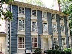 A three story colonial house with blue wood siding in front and brick walls on the sides.