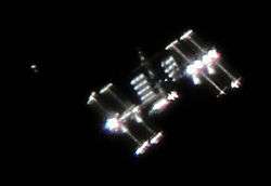 A fuzzy image of the ISS set against a black background, with a smaller, cylindrical object visible to the left of the station.