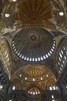 Vertical interior image of the long vaulted ceiling of the nave of Hagia Sophia showing the central ribbed dome with a ring of windows at its base, four pendentives between the four large arches supporting that main dome, two large semi-domes filling the near and far arches (with the other two arches being filled by flat walls with windows, and smaller niche semi-domes in the far large semi-dome