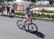 A road racing cyclist in a white jersey with red trim atop a white bicycle, looking behind him out of the frame. Spectators are visible on the roadside behind a barricade.