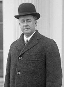 Cleanshaven man with drooping eyelids, aged about 40. He is wearing a black bowler hat, white shirt, tie and dark overcoat