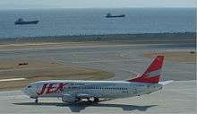A Boeing 737-400 aircraft taxiing on the tarmac, with a seaview of two vessels in the water