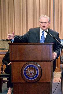 Clean-shaven man in his 60s, with gray hair, wearing glasses, dressed in a dark suit and blue tie, speaking from behind a dark, varnished wooden lectern, with his right arm outstretched.  The front of the lectern is emblazoned with the Great Seal of the United States.