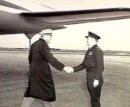 Three-quarter shot of two men at airport shaking hands, one in civilian clothes with dark overcoat and light-coloured hat, the other in military uniform with peaked cap and pilot's wings on battle jacket. An aircraft's tail plane is above and behind the two men.