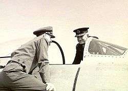 Two men in military uniforms with peaked caps on opposite sides of a military aircraft cockpit with open canopy