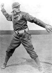 A black-and-white picture of a man in a blue baseball uniform and a white hat winds up to throw a baseball