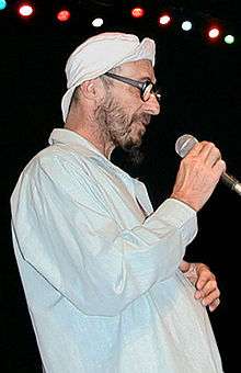 A 55-year-old man is shown in right profile. He wears a light coloured shirt and a white cap with its front rim folded back. He has a beard and wears glasses. He holds a microphone in his right hand and rests his left hand on his lower chest area. There are stage lights over head.