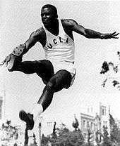 Athlete in UCLA track uniform at the apex of a jump, with legs lunging forward, against a background of an academic building.