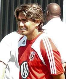 A Hispanic soccer player with shiny brown hair smiles and faces left. He is wearing a red jersey with white and black details and a VW logo.