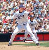 A man in a white baseball uniform with navy blue trim and a navy blue baseball cap throws a baseball from a dirt mound in a stadium with his right hand. His uniform reads "Padres" across the chest in navy blue script trimmed with khaki, and his cap features an interlocked "SD" in white block print. He is wearing a black baseball glove on his left hand.