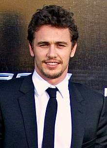 A head shot of James Franco, a caucasian male in his late-20s with dark hair, looking into the camera and smiles. He wears a black suit with a white shirt and black tie, and stands behind a black background.