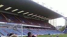 A two-tiered cantilever football stand. The lower tier has light blue seats with some claret seats which spell the word "Clarets". The upper has all claret seating with light blue seats spelling "Burnley".