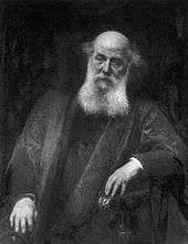 An elderly man wearing dark robes, sitting in a chair with his left arm on the armrest; the top of his head is bald and he has a long white beard