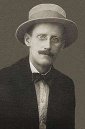 A black-and-white photographic portrait of a mustachioed man with glasses in a brimmed hat.