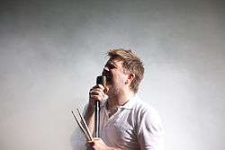 A man sings to a microphone while holding drum sticks.