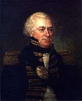 A man with white, receding hair in a high collared military coat with gold accents over a white shirt