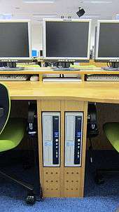A close-up of the space between two student terminals. There are two computers embedded into the desk, and flat-screen monitors sit on top of the desk. The students' headphones are hanging on hooks below the desk.