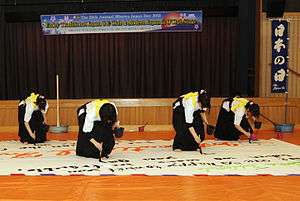 Four female Japanese high school students wearing hakama write calligraphy on a large sheet of paper fixed to the floor. The sheet is around five meters wide and three meters high.