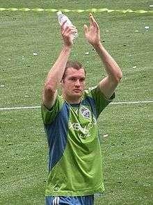 The head and torso of a young man, wearing a green and blue top and blue shorts, standing on a grass field. He has his arms raised above his head in what appears to be a clapping motion.