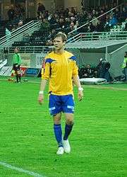 A brown-haired Caucasian American is standing in the center of a grass field, his body facing the camera and his head turned away, and wearing the blue and yellow soccer uniform of his former Norwegian club, Alta.