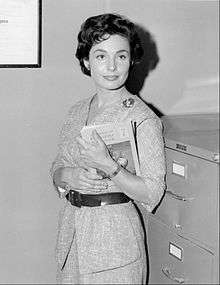 A woman holds a clipboard while standing next to a filing cabinet.