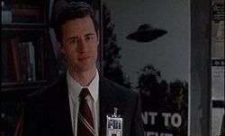 A man is dressed in a white shirt and black suit with a red and cream tie. An FBI badge is clipped on. Behind the man, there is a large poster featuring an UFO.