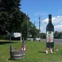 A grassy field along a road decorated with a black cardboard wine bottle and a handful of small flags, with a white wooden sign and flagpole in the background.