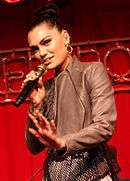 A Caucasian female with her hair in a bun, large gold hoop earrings, and a chocolate-colored animal skin jacket sings into a microphone with her hand flat simulating a pushing motion. She stands in front of a red background and only appears from the waist up.
